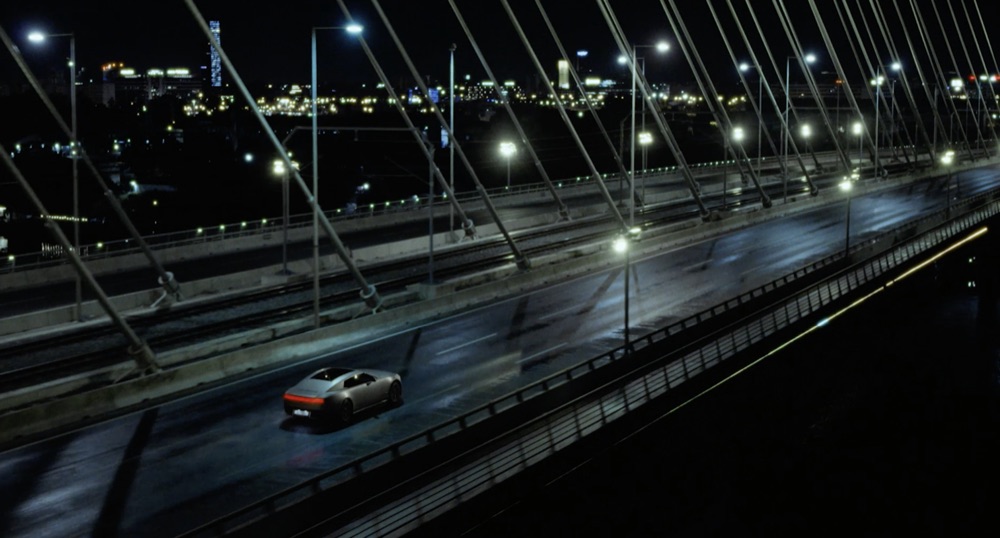 Reference film still: aerial view of a car crossing a floodlit bridge at night